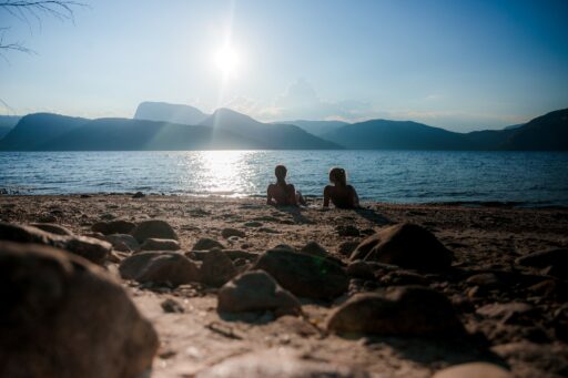 Two people sit on a rocky beach facing a calm lake with distant mountains under a bright sky and sun reflection on the water.