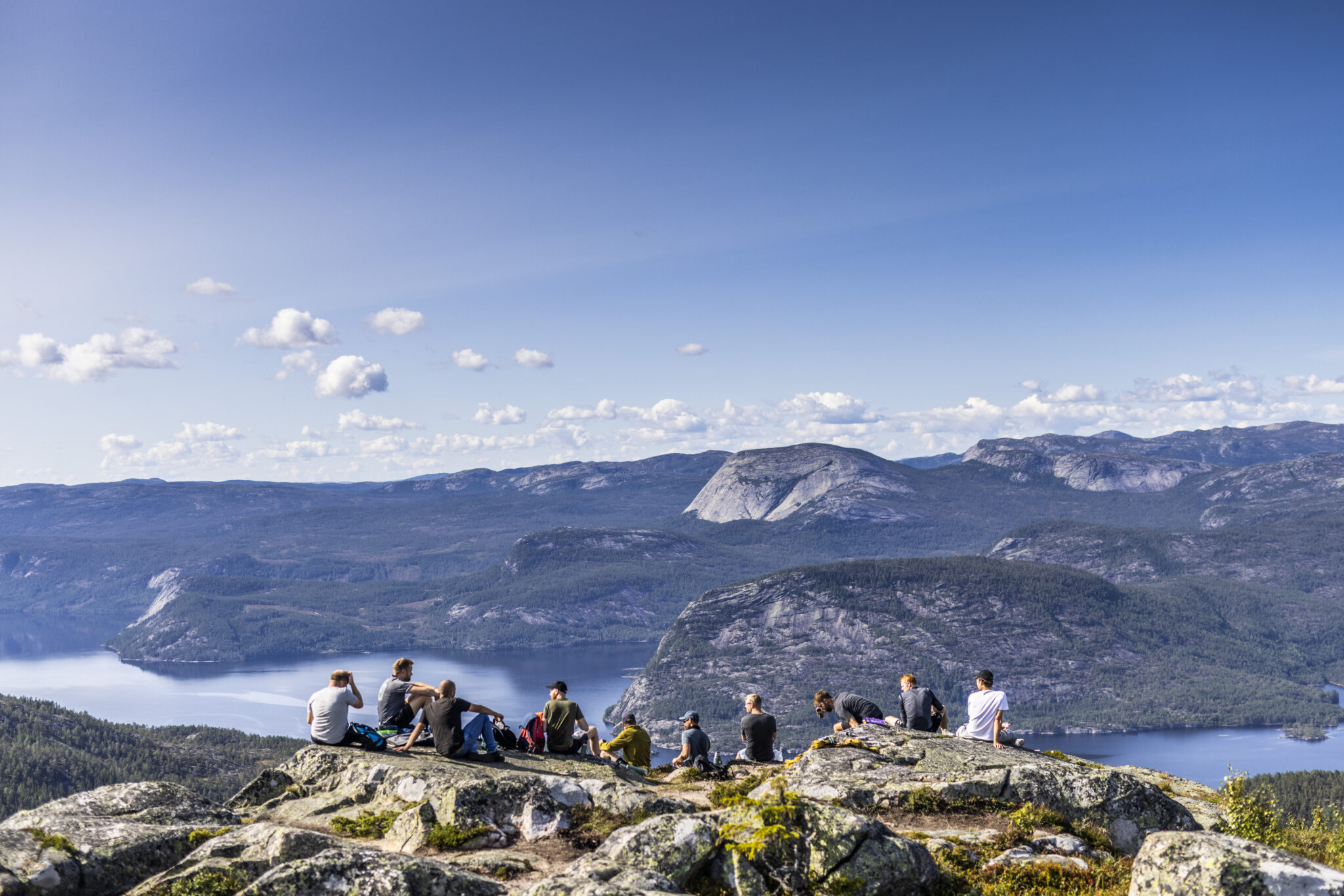 Group of hikers sitting on a rocky overlook, mountains and a fjord below under a clear blue sky