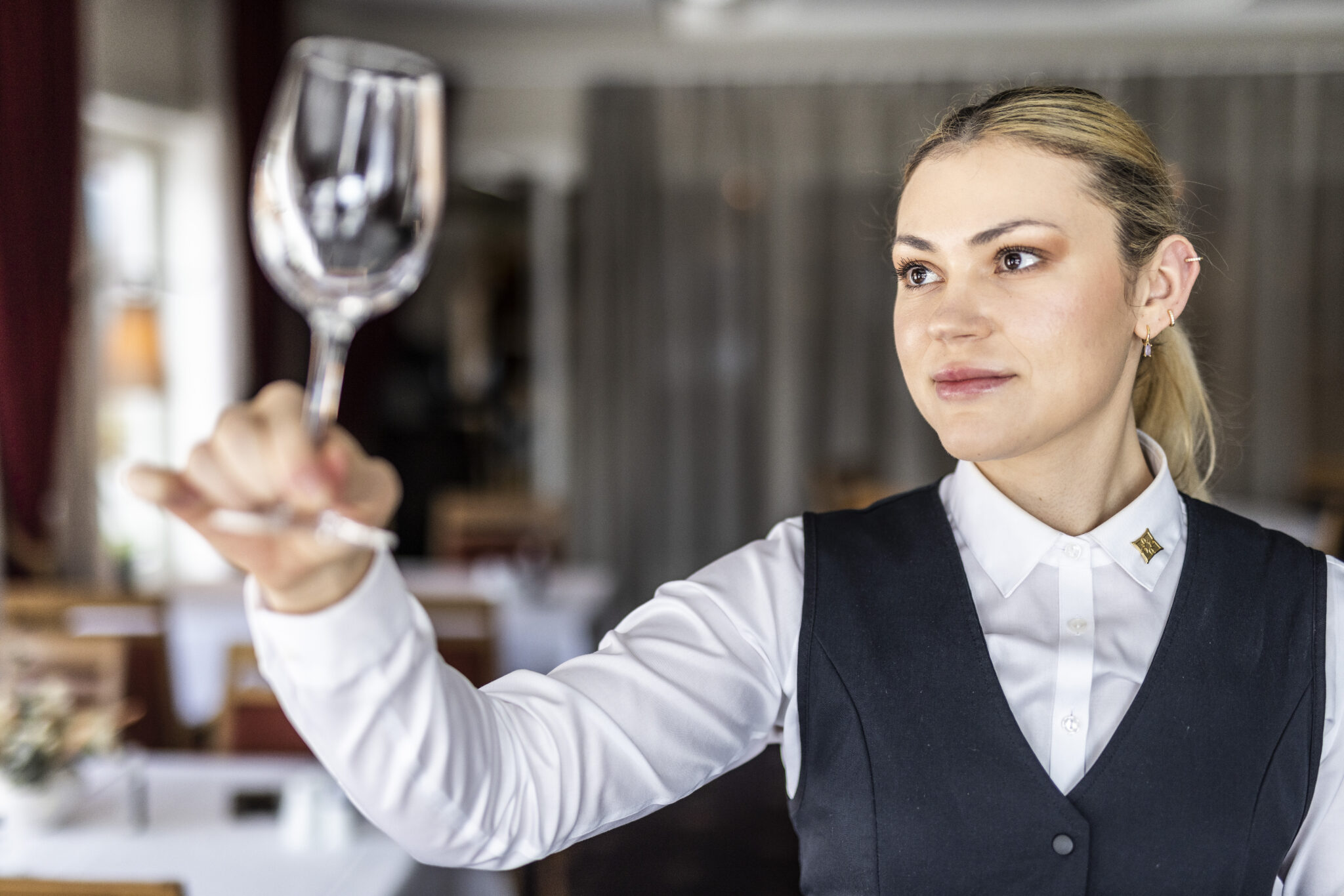 Female server in a white shirt and black vest, holding a wine glass up as if to toast inside a restaurant.