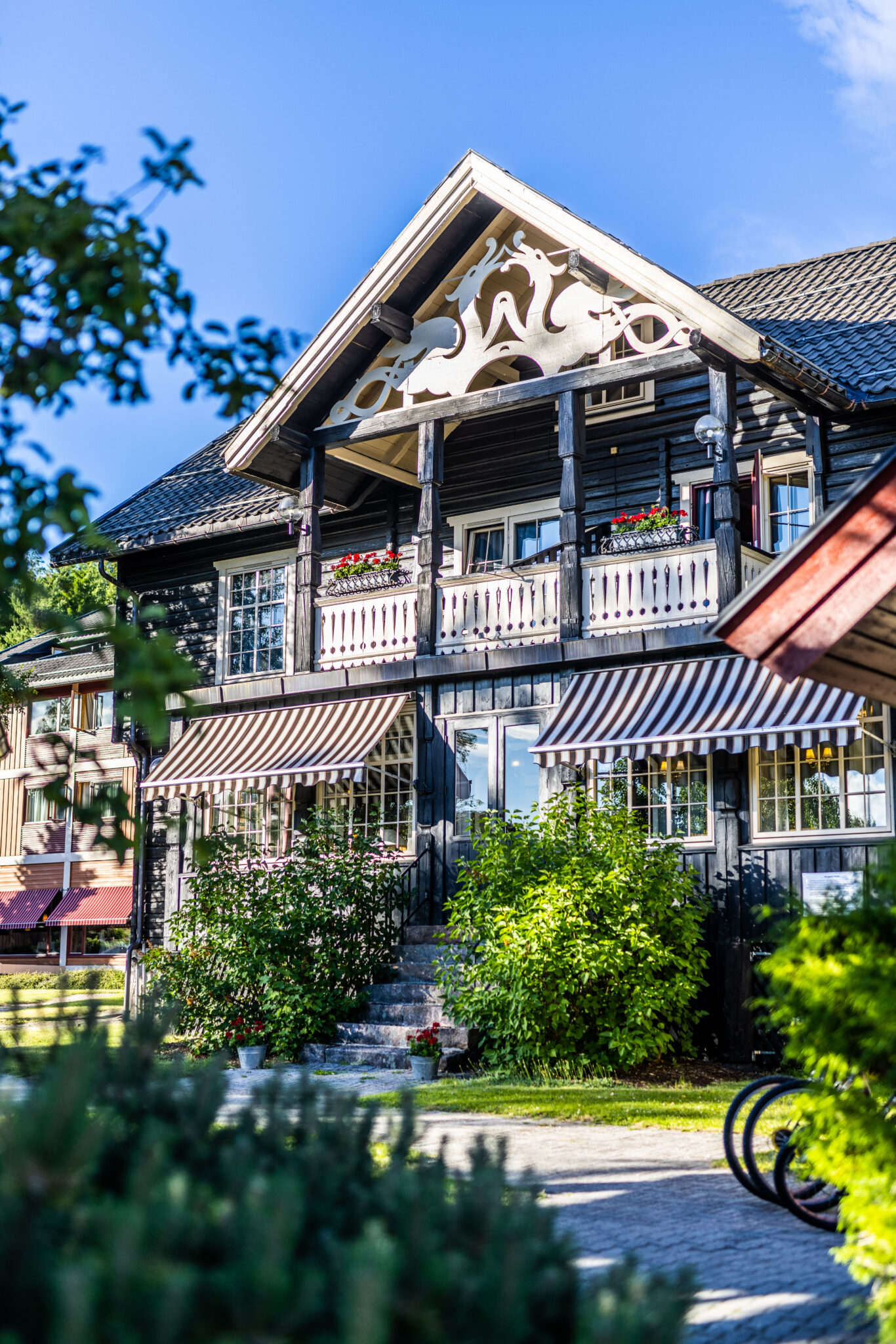 Two-story wooden building with ornate white gable trim, black facade, striped awnings, and flower boxes on a sunny day.