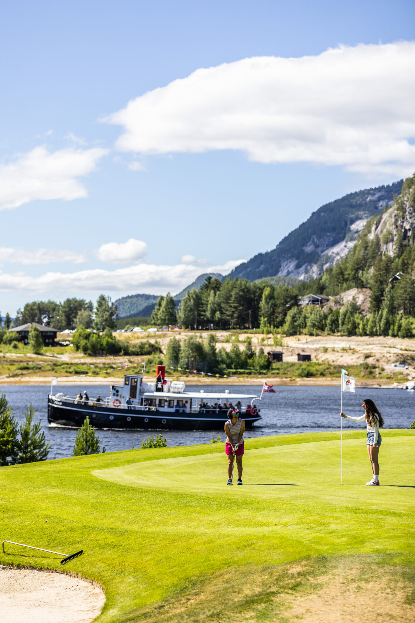 Two people on a bright putting green by a lake, with a boat on the water and forested hills in the background.