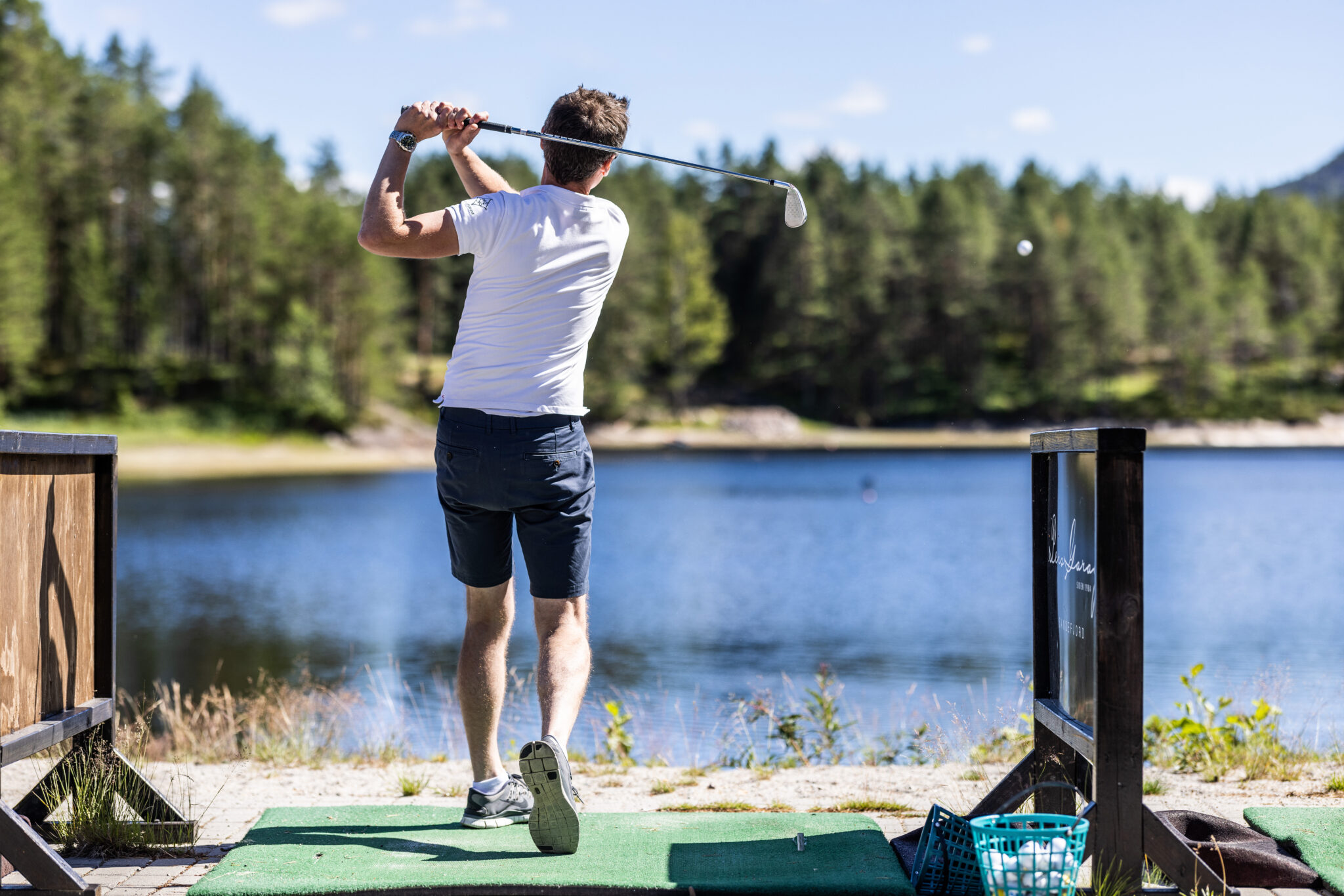 Man in a white shirt and dark shorts swings a golf club on a lakeside wooden deck.