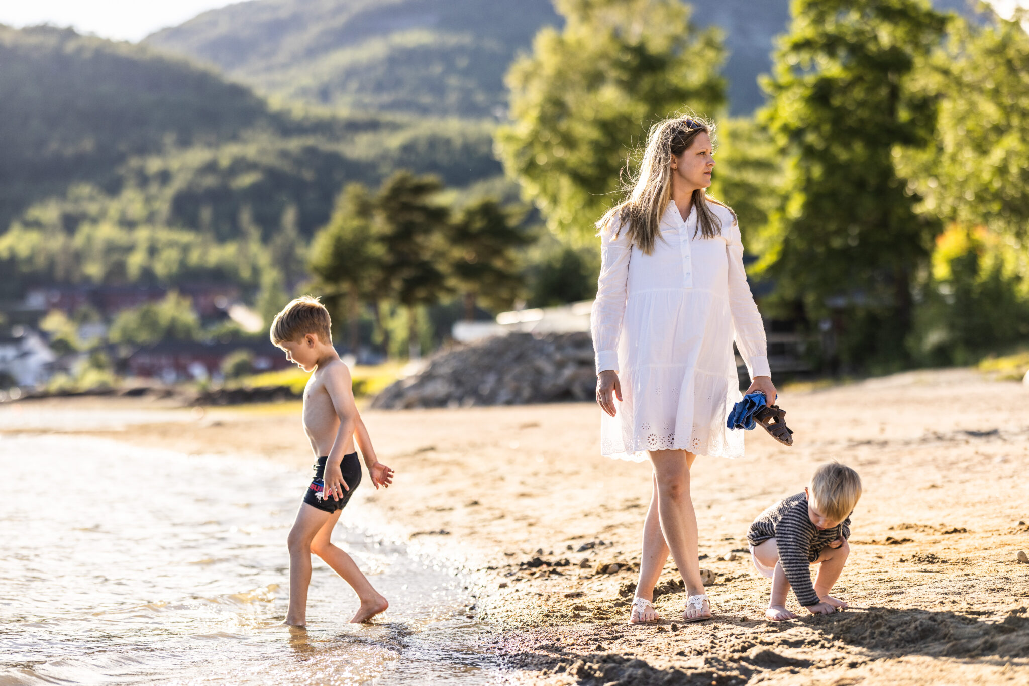 Mother in a white dress stands on a sunny beach with children playing at the water's edge, hills in the distance.