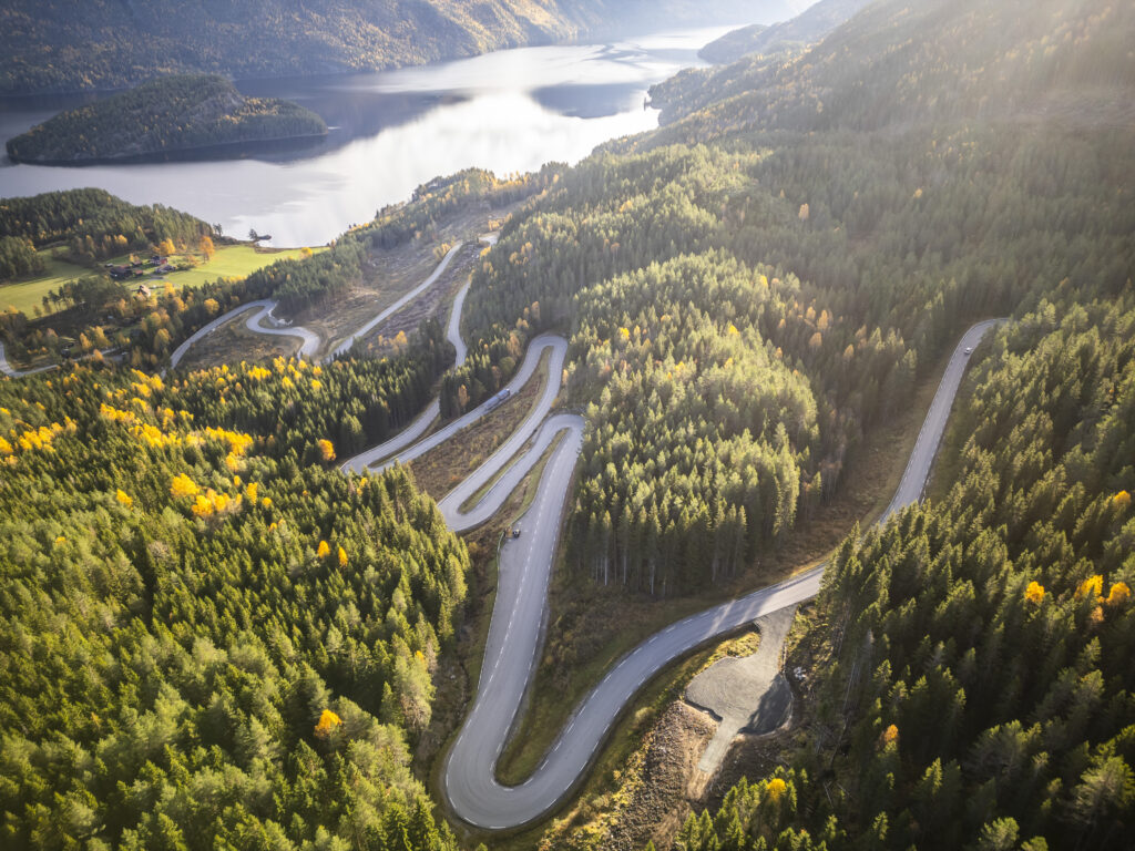 Aerial view of a winding mountain road cutting through a dense forest with autumn colors and a lake in the distance.