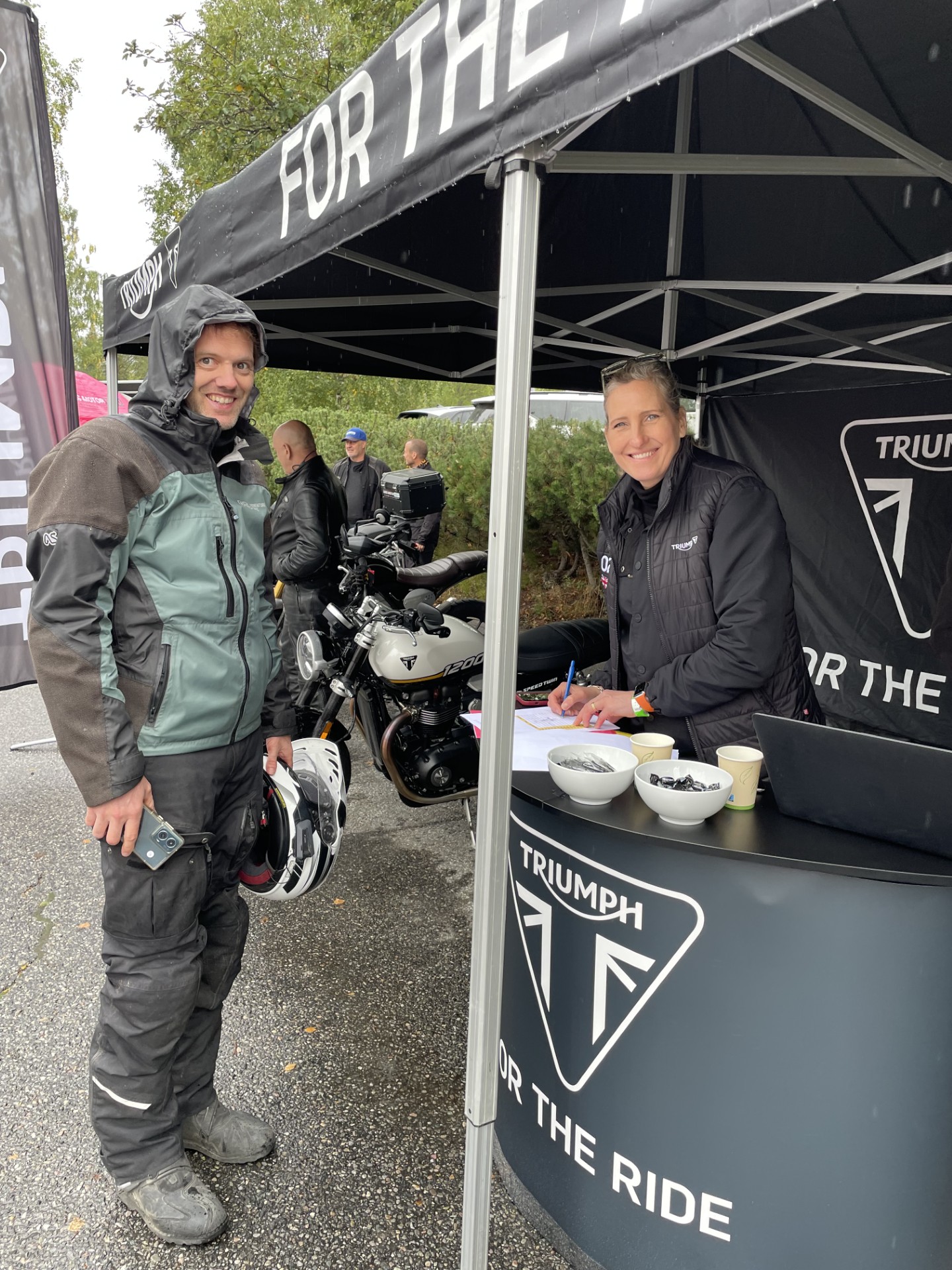 Motorcyclist in rain gear holding a helmet chats with a staff member at a Triumph booth; a Triumph motorcycle is visible behind them in the tent area.