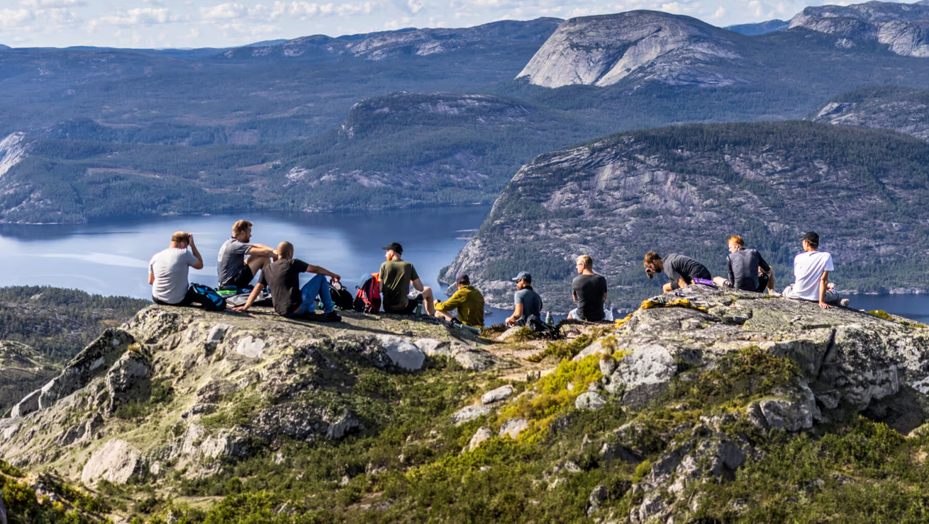 A group of hikers sits on a rocky overlook, looking out over a blue fjord and rugged mountains.