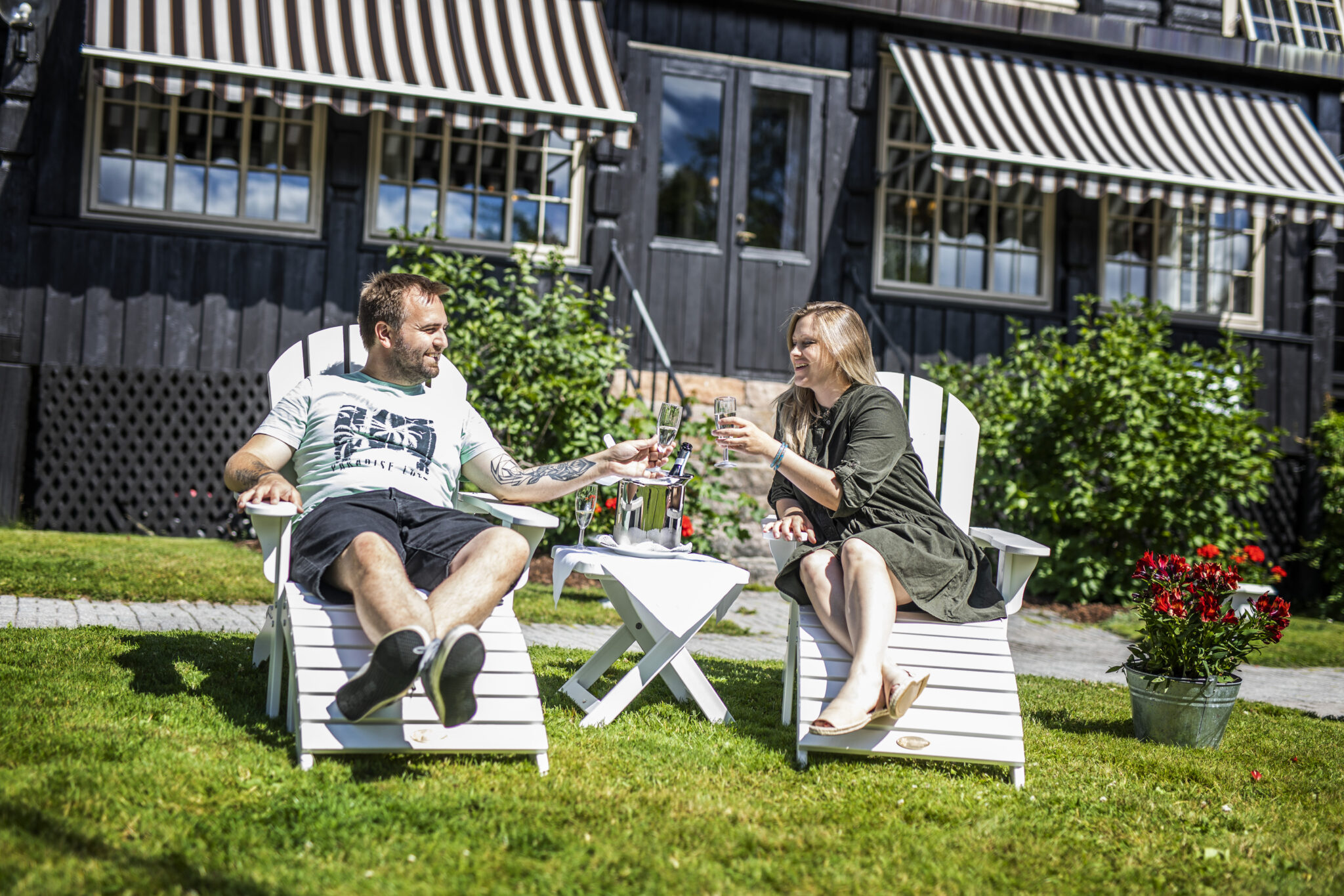 Man and woman toasting with champagne in white Adirondack chairs in a sunny garden in front of a dark wooden house with striped awnings.