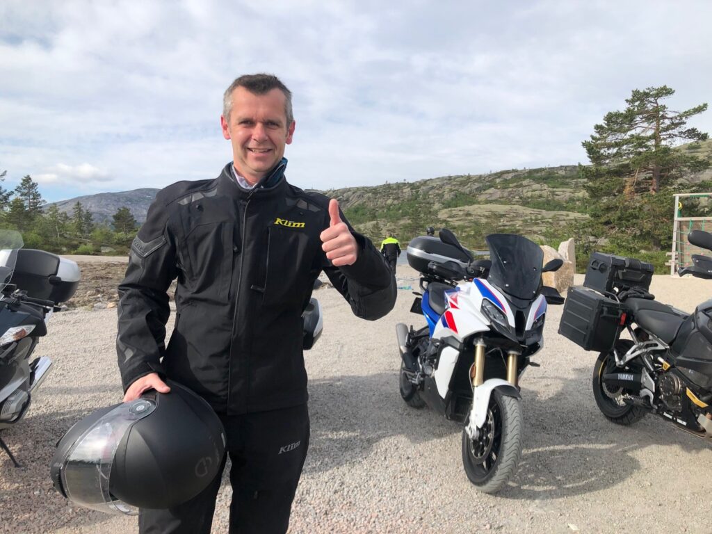 Smiling man in a black Klim riding jacket holding a helmet, giving a thumbs up, with touring motorcycles parked behind him and rocky hills in the distance.
