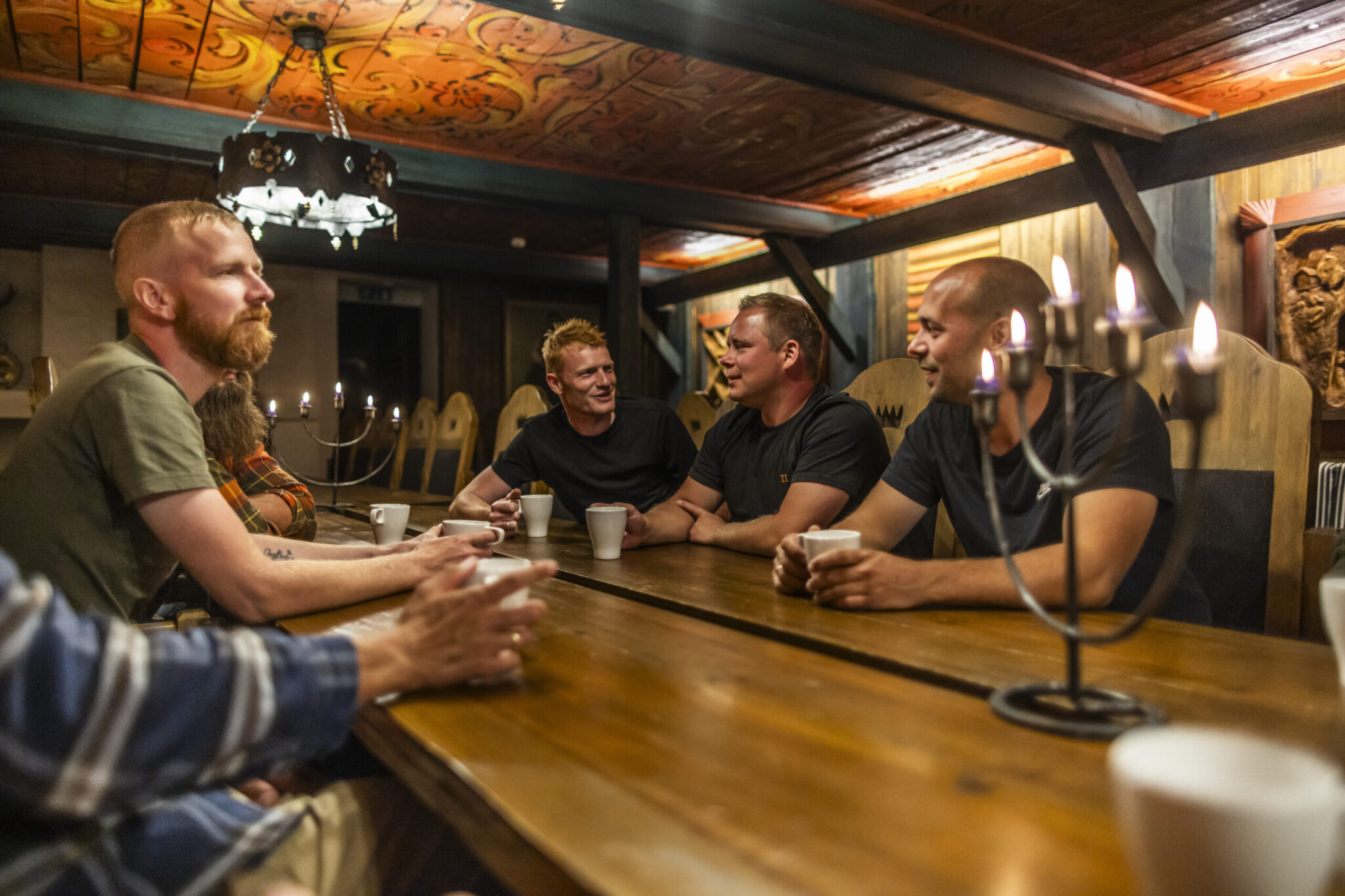 Group of men sitting around a wooden dining table in a rustic room, chatting over white mugs beneath warm candlelight.