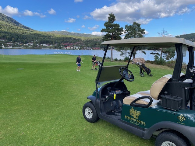 Golf course beside a lake with pine trees and mountains in the background; a golf cart sits in the foreground.