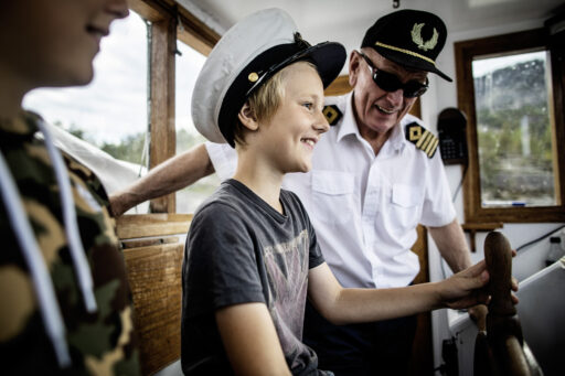 Boy wearing a white captain's hat smiles while a man in a white shirt and cap chats with him inside a wooden boat cabin.