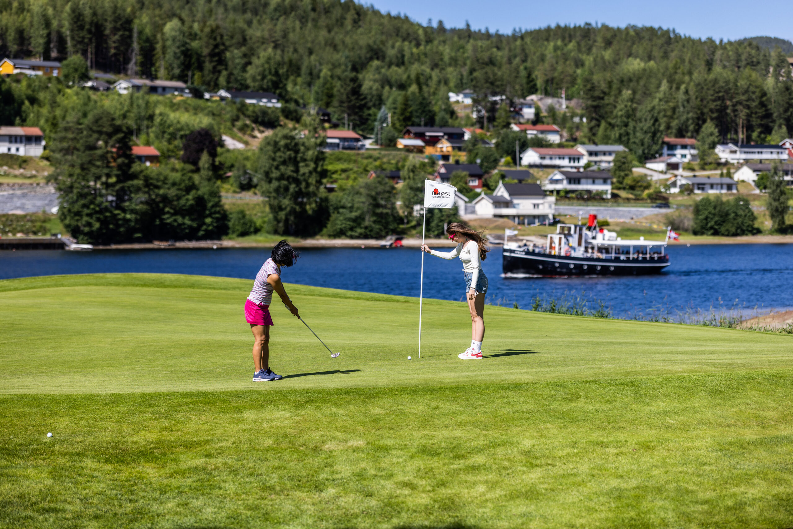Two women golfer in pink shorts and white top near a flag on a sunny lakeside golf course with a boat and houses in the background
