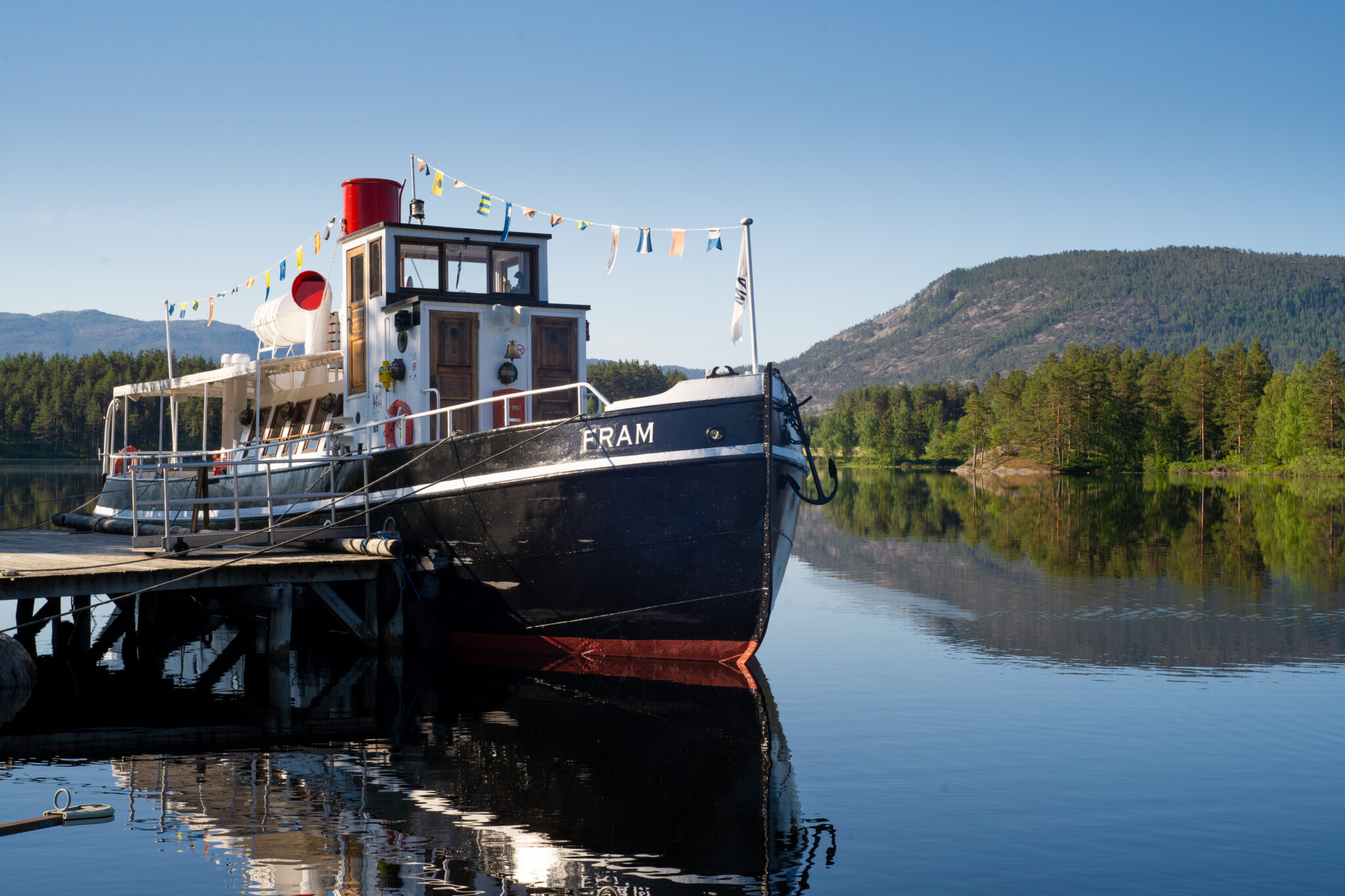 A classic boat docked at a wooden pier on a calm lake, with colorful pennant flags and forested hills in the background.