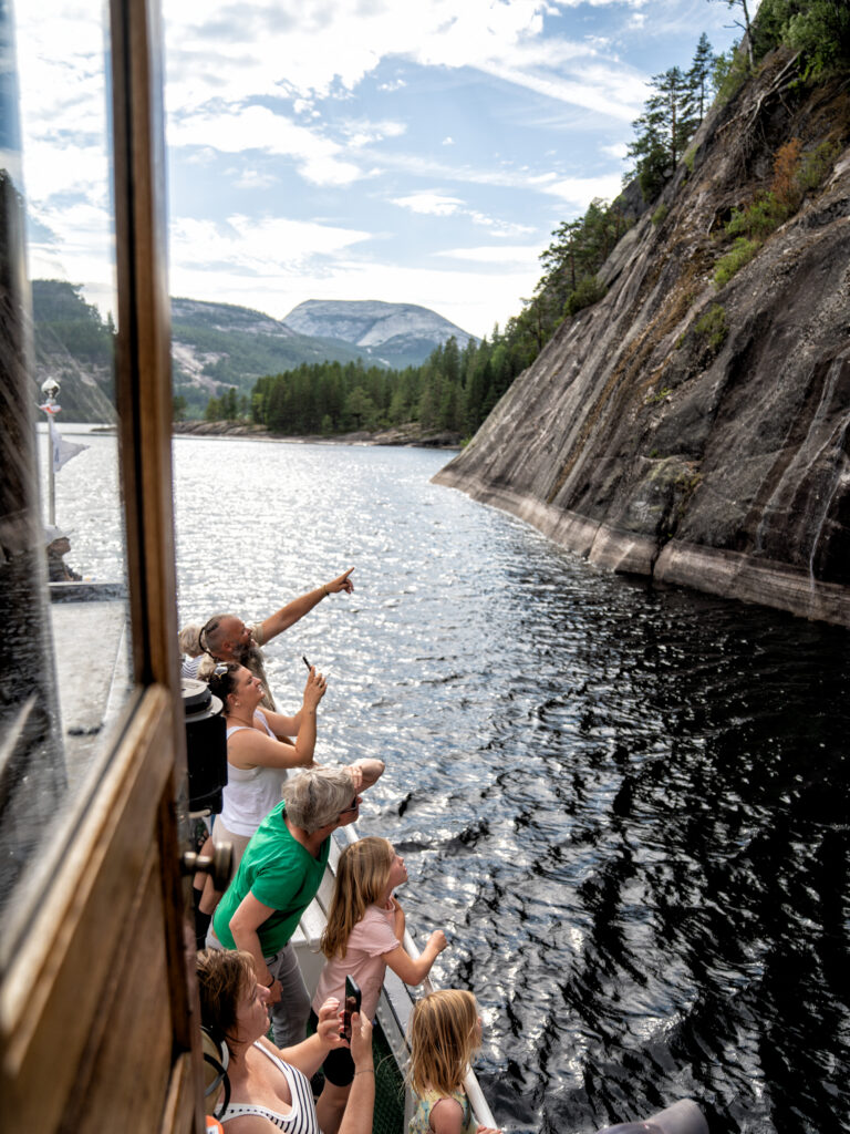 Group of tourists on a boat looking over a rocky cliff at a calm lake with mountains in the background in bright daylight