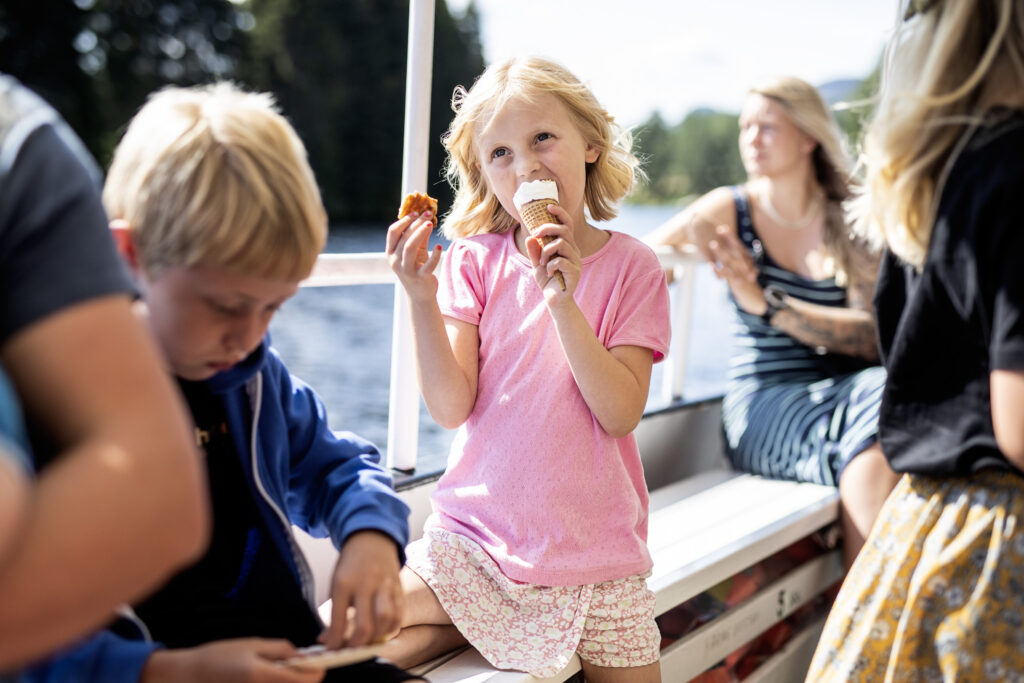 Girl in pink shirt eats an ice cream cone on a boat, with friends nearby on sunny water.