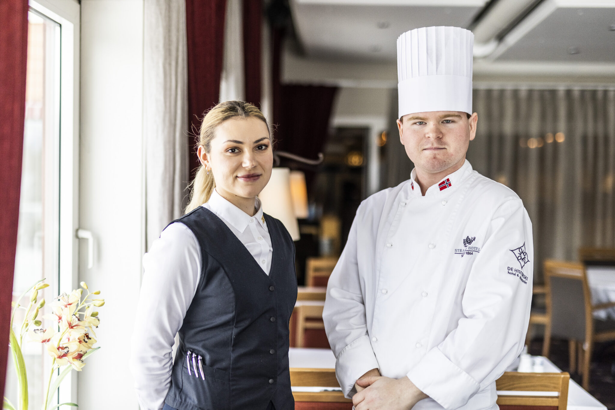 Two restaurant staff members pose beside a window: a female server in a dark vest and white shirt, and a male chef in a white uniform and tall hat.