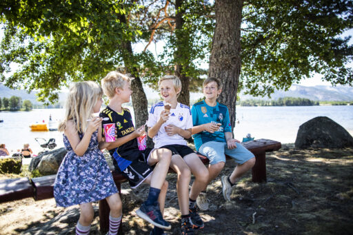 Group of smiling children sitting on a wooden bench by a lakeside under trees, eating ice cream cones.