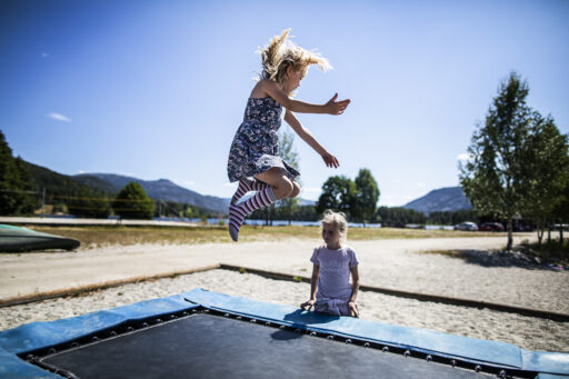 Two young girls play on a blue trampoline outdoors on a sunny day, one mid-jump with hair flying and arms outstretched.