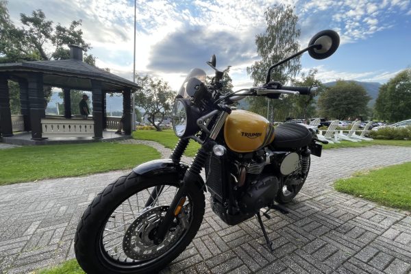 Gold Triumph motorcycle in the foreground with a round headlight, windshield, and black frame parked on a brick path in a park A yellow Triumph motorcycle parked near a gazebo with trees, hills, and parked cars in the background Close-up of a Triumph motorcycle’s front end on a paved plaza, with a park gazebo and mountains visible behind it Motorcycle-focused scene: a yellow Triumph bike on a brick walkway beside a wooden gazebo and grassy park area