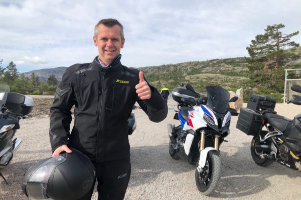 Smiling man in a black Klim riding jacket holding a helmet, giving a thumbs up, with touring motorcycles parked behind him and rocky hills in the distance.
