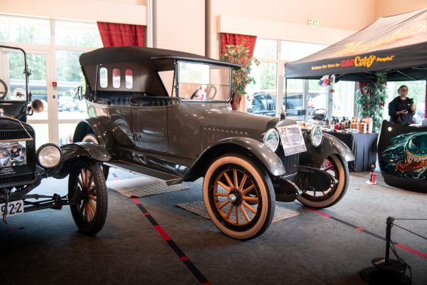Vintage open-top automobile on display indoors at a car show, black/gray body with wooden wheels and tufted interior.