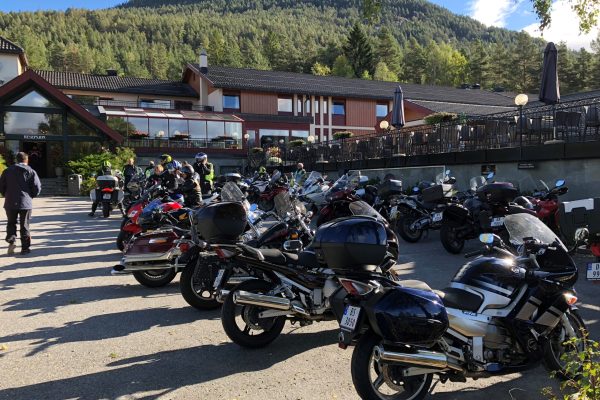 Parked motorcycles fill a lot outside a mountain-view lodge, with riders in gear and a few people walking around.
