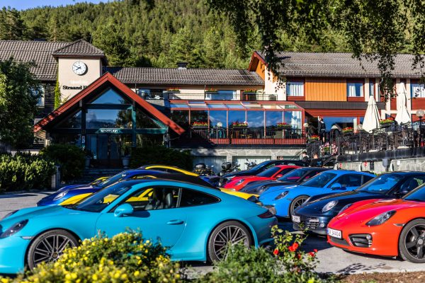 Row of bright blue, yellow, and red sports cars parked in front of a hotel/resort building with a glass atrium, clock tower, and forested hillside behind.